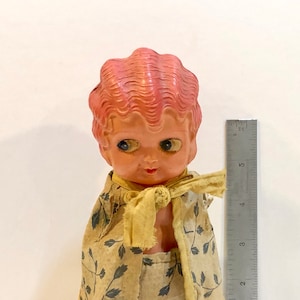 May include: A vintage doll with a pink, wavy hairstyle and large, expressive eyes. The doll is wearing a yellow scarf and a patterned outfit with blue leaf designs. A ruler is placed next to the doll, measuring approximately 5 inches.