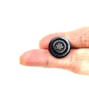 May include: A small, circular black button featuring a recessed center with a raised six-pointed star design. The button has a smooth, dark finish and a distinct border around the central star motif, creating a simple and functional fastener.