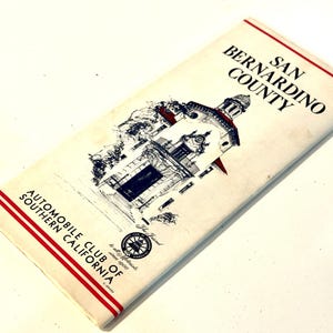 Könnte beinhalten: Eine alte weiße Papierkarte von San Bernardino County mit roten Randlinien und einer schwarzen Tuschezeichnung eines Gebäudes. Der Text lautet SAN BERNARDINO COUNTY und AUTOMOBILE CLUB OF SOUTHERN CALIFORNIA mit einem kleinen runden Logo.
