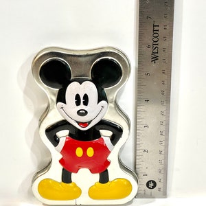 May include: A Mickey Mouse-shaped tin with a silver finish. The cartoon character is depicted in his classic outfit: black body, red shorts, yellow shoes, and white gloves. A ruler is next to the tin, measuring approximately 5 inches.