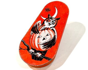 May include: An oval-shaped orange metal tin featuring a graphic illustration of an owl perched on a branch while holding a jack-o'-lantern pumpkin. A white crescent moon is visible above the owl, and the text U.S. METAL TOY MFG. CO. is printed.