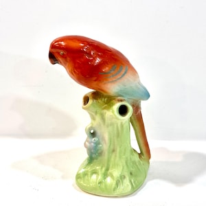 May include: A ceramic parrot figurine perched on a green tree stump. The parrot has vibrant red, orange, and blue plumage. The tree stump is light green with a small cluster of purple berries. The figurine is a decorative item.
