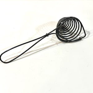 May include: A black metal kitchen utensil with a spiral-shaped head and a long handle. The utensil appears to be a whisk or a similar tool, designed for mixing or aerating ingredients. The handle has a loop at the end.