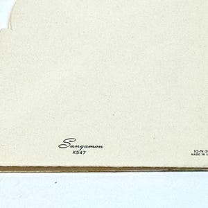 May include: A close-up of a vintage greeting card with a cream-colored background. The card has a decorative edge on the left side. The word "Sangamon" is printed in cursive, along with "K547" and "10-N-300 MADE IN U.S.A."