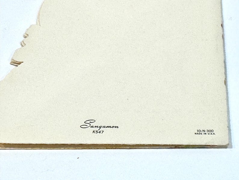 May include: A close-up of a vintage greeting card with a cream-colored background. The card has a decorative edge on the left side. The word "Sangamon" is printed in cursive, along with "K547" and "10-N-300 MADE IN U.S.A."