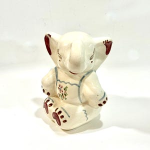 May include: A ceramic elephant figurine in a seated position, painted in off-white with red and blue accents. The elephant wears blue overalls with a floral design on the chest and red details on the ears and feet.