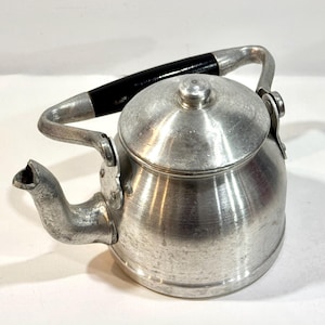 May include: A vintage aluminum tea kettle with a black handle. The kettle has a spout, a lid, and a handle that arches over the top. The kettle is made of metal and has a shiny, reflective surface. The kettle is designed for heating water.