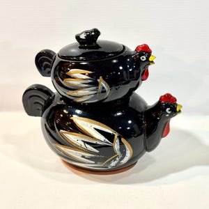 May include: A black ceramic teapot and lid, shaped like a stacked hen and chick. The teapot has a red comb and yellow beak. White and gold leaf-like designs adorn the sides. The lid has a small knob. A unique kitchenware item.