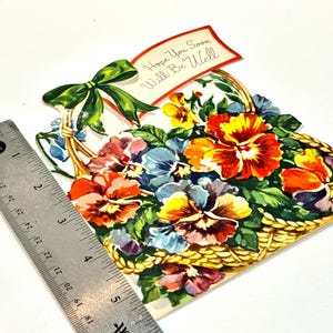 May include: A vintage get-well card shaped like a basket filled with colorful pansies. The card features a green bow and a banner that reads "Hope You Soon Will Be Well". The card is approximately 5 inches wide.