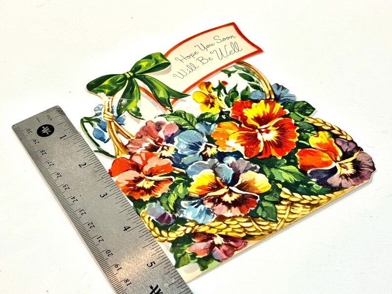 May include: A vintage get-well card shaped like a basket filled with colorful pansies. The card features a green bow and a banner that reads "Hope You Soon Will Be Well". The card is approximately 5 inches wide.