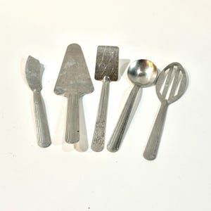 Puede incluir: Un conjunto de cinco utensilios de servir plateados vintage. Incluye un cuchillo de mantequilla, una espátula para tartas, una espátula, una cuchara y una cuchara ranurada. Los utensilios están dispuestos sobre una superficie blanca, mostrando su diseño simple y funcional.