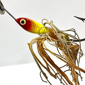 May include: A fishing lure with a red and yellow painted body, a silver spinner blade, and a large hook. The lure has a cluster of brown and tan fibers extending from the body.