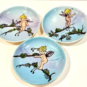 Könnte beinhalten: Drei Vintage-Keramikplatten mit hellblauem, gesprenkeltem Hintergrund. Jede Platte zeigt eine skurrile Illustration einer Fee mit gelbem Haar und lila Flügeln, die auf einem Ast sitzt. Die Teller sind wahrscheinlich dekorativ.