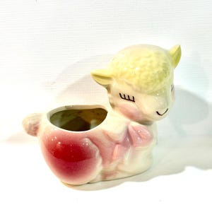 Könnte beinhalten: Ein Keramik-Pflanzgefäß in Form eines liegenden Lamms mit gelbem, strukturiertem Kopf, rosa Wangen und rotem Körper. Das Lamm hat geschlossene Augen mit langen schwarzen Wimpern und ein kleines Lächeln. Die Öffnung für eine Pflanze ist am Rücken.