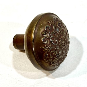 May include: Antique brass door knob with an ornate floral design. The knob has a rounded shape with a detailed pattern on the front. The finish is a warm, aged brass color, suggesting a vintage aesthetic. The knob is designed for door hardware.