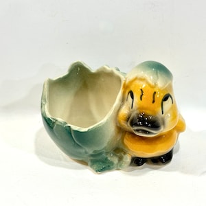 May include: A ceramic duckling planter, featuring a duck emerging from a cracked eggshell. The duckling has a yellow body, black beak, and a blue-green head. The eggshell is a matching blue-green color.