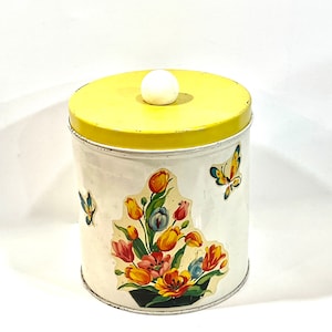Puede incluir: Un bote de metal blanco vintage con tapa amarilla y pomo blanco. El bote está decorado con un ramo de flores y calcomanías de mariposas. El bote es cilíndrico y tiene un acabado envejecido.