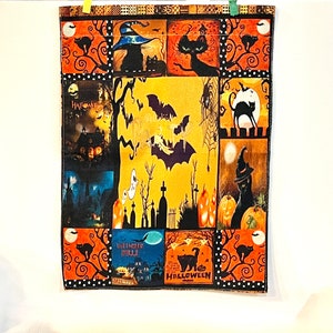 May include: A rectangular fabric wall hanging featuring a collage of Halloween-themed illustrations including black cats, pumpkins, bats, ghosts, a haunted house, and a graveyard. The design uses a colour palette of orange, black, and yellow.