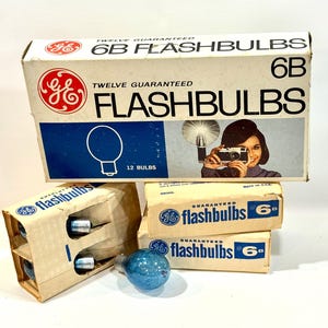 May include: Vintage GE 6B Flashbulbs boxes. The boxes are white and tan with the GE logo and the words "Flashbulbs" and "Twelve Guaranteed" printed on them. The boxes contain flashbulbs and a blue glass bulb.