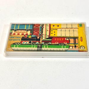 Può includere: Una custodia rettangolare in plastica contenente un puzzle con una colorata illustrazione di un treno. Il treno è rosso, nero e verde con la parola POST stampata sul lato. Lo sfondo presenta forme geometriche gialle, blu e rosse.