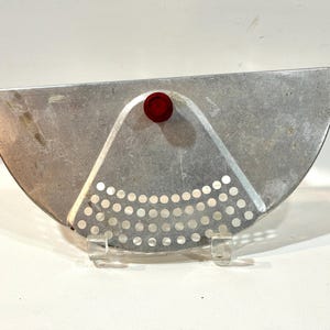 May include: A semi-circular aluminum pot strainer featuring a flat edge and a curved perforated section with small circular holes. The kitchen tool includes a small, round, dark red knob centered at the top for handling and grip.