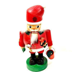 May include: A wooden nutcracker figure wearing a red hat and jacket with white trim. It features a white beard, black boots, and a green base. The figure holds a small hammer and a red bucket, with a small orange star-shaped ornament attached.