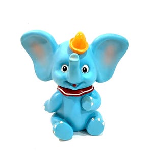 May include: A light blue elephant figurine with large ears, a yellow hat perched on its head, and a red and white collar around its neck. The elephant has black eyes, a small red mouth, and white painted details on its feet and toes.