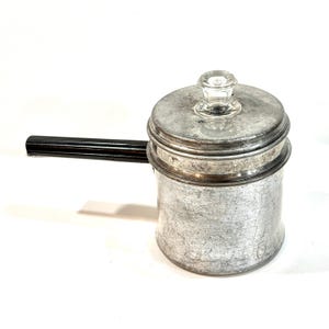 Może przedstawiać: Wintage aluminiowy dzbanek do kawy z czarnym uchwytem i przezroczystym szklanym pokrętłem na pokrywce. Dzbanek ma cylindryczny korpus i pokrywkę. Uchwyt wykonany jest z ciemnego, prawdopodobnie żaroodpornego materiału.