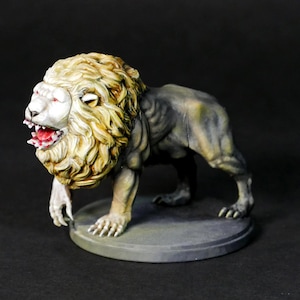 Op de afbeelding: Een geschilderd miniatuurbeeldje van een leeuw met een wit gezicht en een grommende uitdrukking. De leeuw staat op een ronde basis.