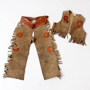 May include: A brown leather cowboy chaps and waistcoat set with fringe detailing and decorative stitching. The chaps have a belt with metal accents and the waistcoat has a fringed collar.