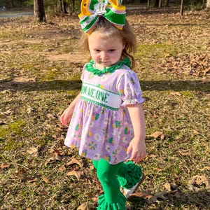 May include: A young girl wearing a purple dress with a green collar and the text "SHOW ME ONE". The dress has a pattern of colorful shapes. She is also wearing green leggings with ruffled cuffs, a large bow in her hair, and white shoes.