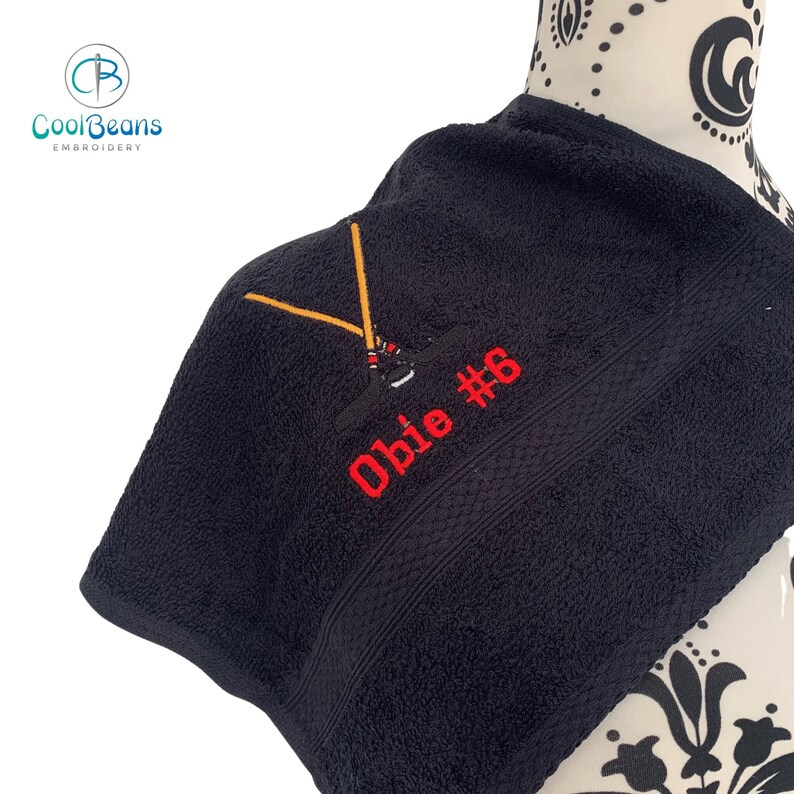 May include: Black towel with embroidered hockey sticks and the text "Obie #6" in red thread.