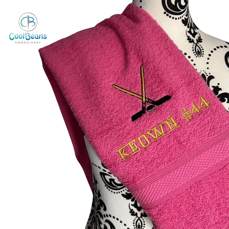 May include: A pink towel with gold embroidery of two hockey sticks crossed with a puck and the text "KEOWN #44".