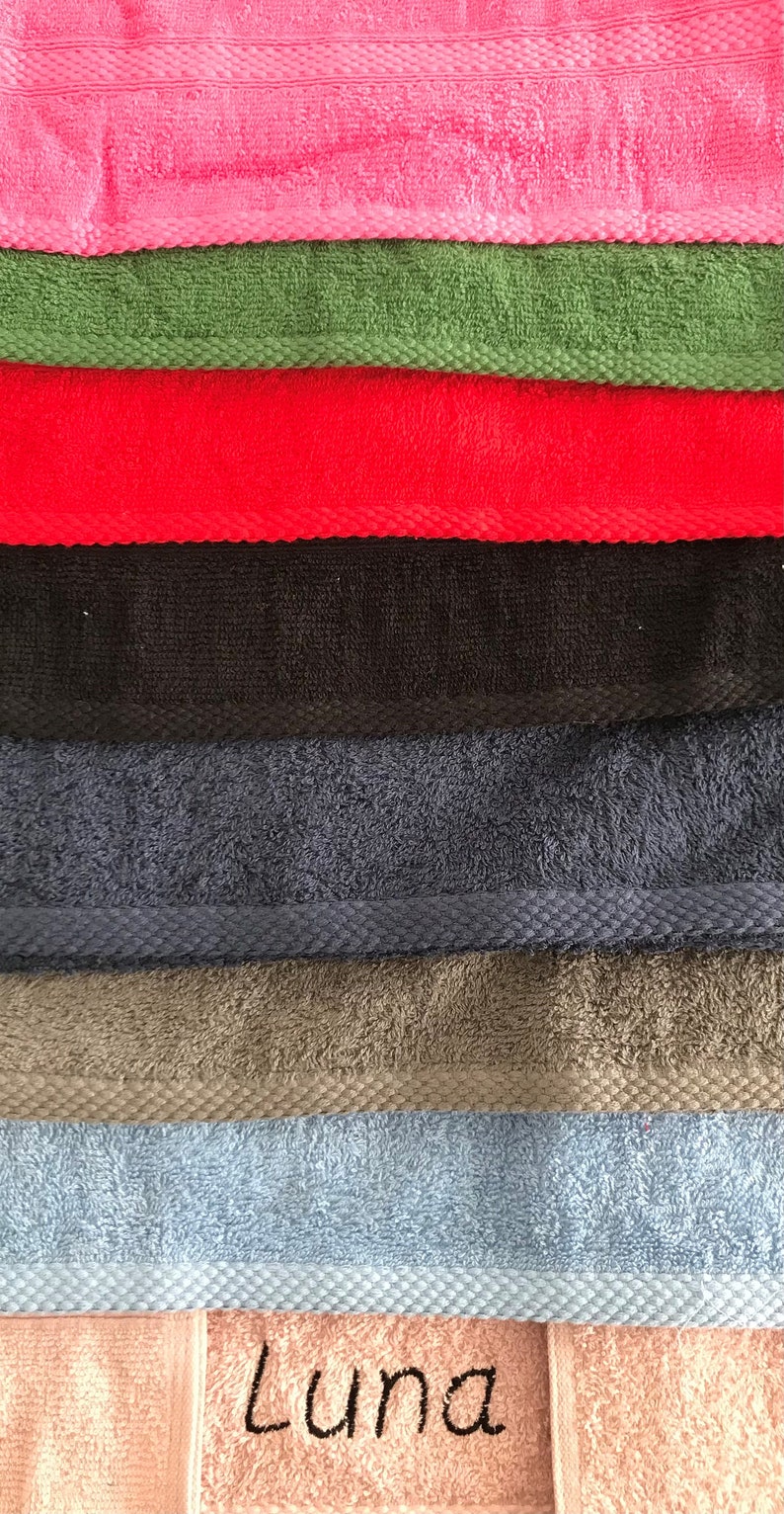 May include: A stack of eight towels in various colors, including pink, green, red, black, blue, gray, light blue, and peach. The peach towel has the name "Luna" embroidered on it.