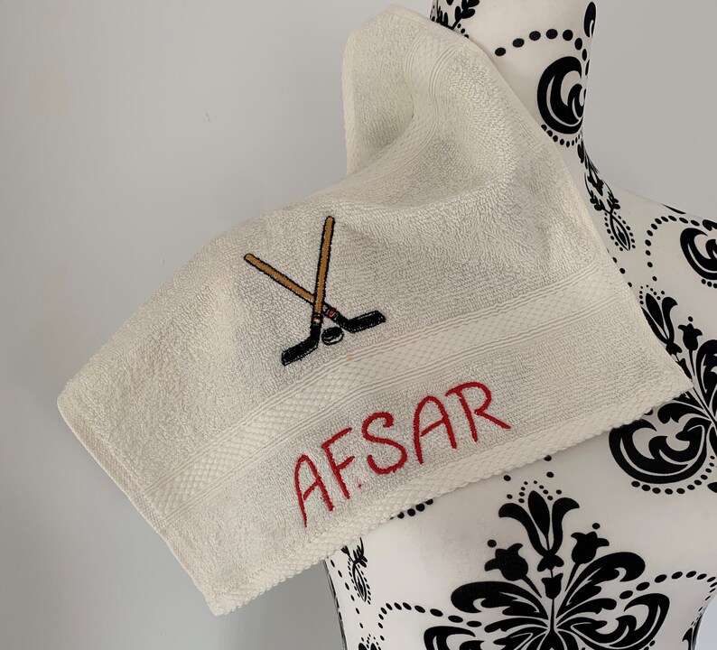 May include: White towel with red embroidery of the name "AFSAR" and a pair of crossed hockey sticks with a puck.