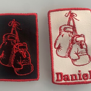 May include: Two red embroidered patches featuring boxing gloves hanging from a string. One patch is black with red stitching and the other is white with red stitching. The white patch has the name "Daniel" embroidered on it.