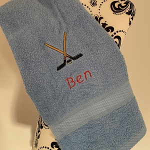 May include: A light blue towel with a personalized embroidered design featuring crossed hockey sticks and the name "Ben".