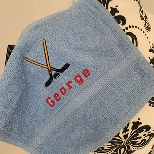 May include: A light blue towel with a red embroidered name "George" and a pair of crossed hockey sticks.