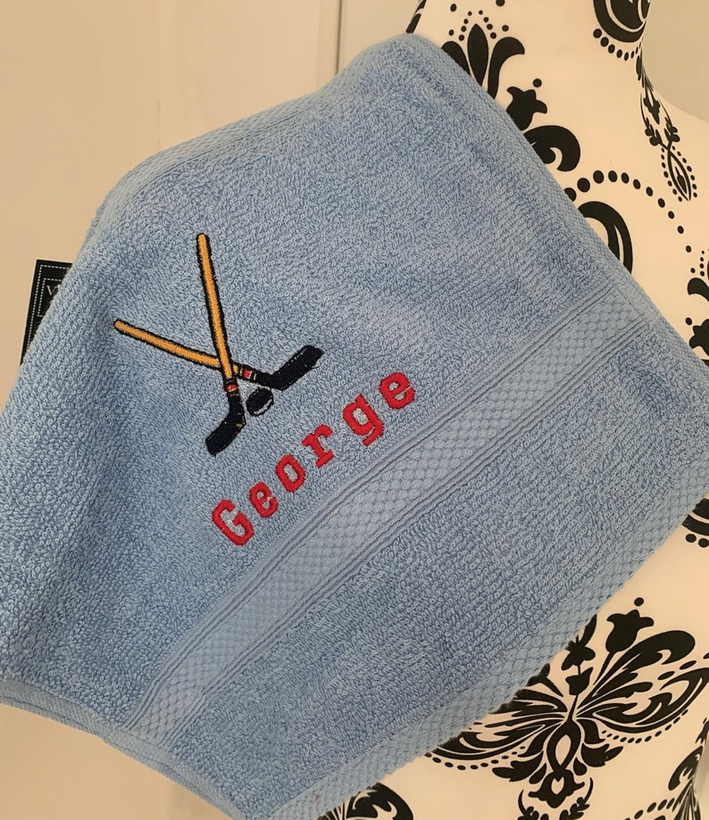 May include: A light blue towel with a red embroidered name "George" and a pair of crossed hockey sticks.