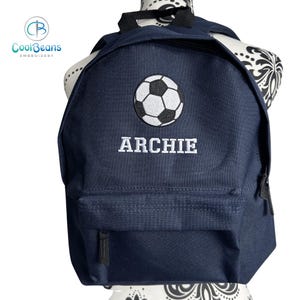 May include: A navy blue backpack featuring a soccer ball emblem and the name "ARCHIE" embroidered in white. It has a front pocket and adjustable straps. The CoolBeans Embroidery logo is visible on the left.