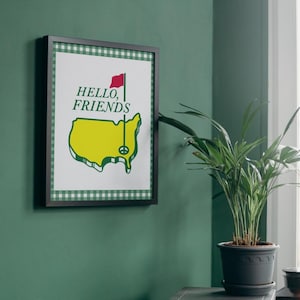 May include: A framed print with a green and white gingham border. The print features a yellow outline of the United States with a peace sign in the centre. The text "HELLO, FRIENDS" is above the outline. A red flag is at the top of a golf flagpole.