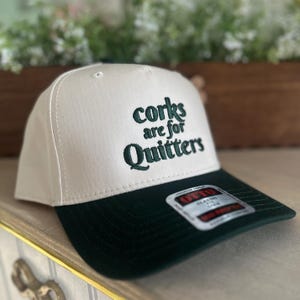May include: A white and dark green baseball cap with the text "corks are for Quitters" embroidered on the front. The cap has a classic fit and a structured design, suitable for casual wear.