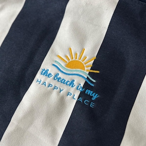 Puede incluir: Tela a rayas blancas y azul marino con texto bordado que dice "the beach is my happy place" y un sol amarillo con olas azules.