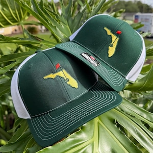 May include: Two dark green and white trucker hats featuring an embroidered yellow map of Florida with a red golf flag pin. The hats have a structured front panel, a flat brim with decorative stitching, and a white mesh back for ventilation.