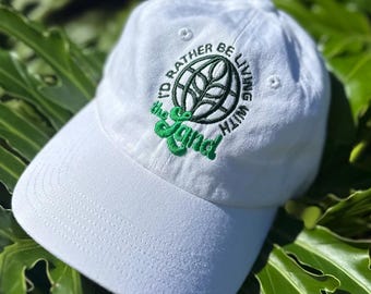 Könnte beinhalten: Weiße Baseballkappe mit grüner Stickerei, auf der "I'd rather be living with the land" steht. Die Stickerei zeigt ein grünes Blatt in einem schwarzen Kreis.