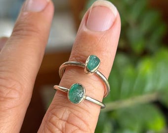 May include: Two silver rings stacked on a finger, each featuring a small, raw, oval-shaped green emerald gemstone set in a simple metal bezel. The bands are thin and polished, showcasing a minimalist design with natural, uncut green stones.