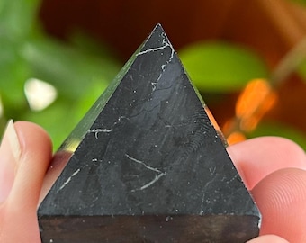 May include: A small, solid black pyramid object with a smooth, matte finish and subtle, thin white veining patterns running across its triangular faces. The geometric shape features sharp, clean edges and a flat, square base.