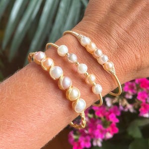 May include: Three gold-toned bracelets adorned with pearls of varying sizes and colors, including white and peach hues. The bracelets are displayed on a wrist, with a backdrop of green foliage and vibrant pink flowers.