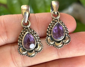 May include: Two silver pendants featuring a central pear-shaped faceted purple amethyst gemstone. Each pendant has a decorative border with small silver beads and a smooth metal base, topped with a silver bail for attachment to a chain or cord.