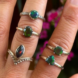Pode incluir: Uma coleção de anéis de prata, cada um com uma pedra preciosa de opala verde escura. Os anéis apresentam vários designs, incluindo pedras redondas, ovais e triangulares. Os anéis são exibidos em dedos contra um fundo desfocado.